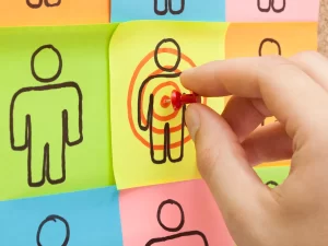 Nine figures drawn on colored paper with a hand putting a thumbtack on an identified target person.